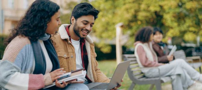 Happy students sitting on bench, looking at laptop and discussing their project together